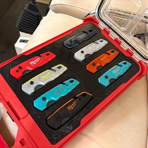 Peut inclure: Un coffret rouge Milwaukee Packout contenant huit couteaux utilitaires pliants de différentes couleurs, dont rouge, orange, jaune, bleu et noir, tous arborant le logo Milwaukee, organisés dans un insert en mousse noire découpé sur mesure.