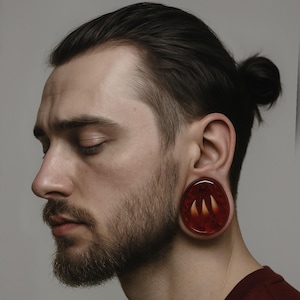 May include: A close-up of a person wearing a dark red ear gauge. The gauge is circular, with a transparent red base and a design of three yellow-orange claw-like shapes. The person has a beard and dark hair pulled back.