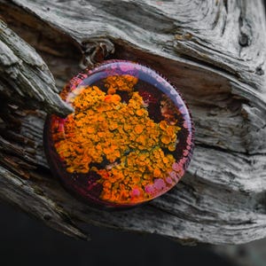 Op de afbeelding: Een ronde hars hanger met een abstract ontwerp in oranje, gele en roze tinten. De kleuren zijn ingekapseld in heldere hars. De hanger is geplaatst tegen een achtergrond van verweerd, grijs hout, wat een opvallend contrast creëert.