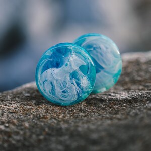 Op de afbeelding: Twee ronde blauwe en witte gemarmerde oorpluggen. De pluggen hebben een draaiende witte en blauwe print.