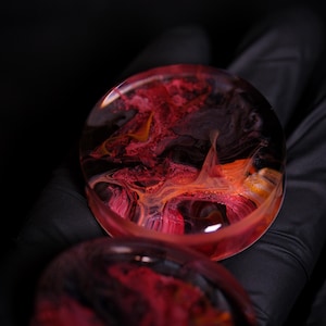 May include: Two round, clear resin ear gauges with a swirling red, orange, and black pattern.