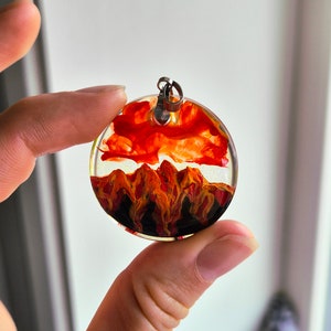 May include: A round resin pendant with a red, orange, and black abstract design. The pendant has a silver bail for attaching to a necklace.