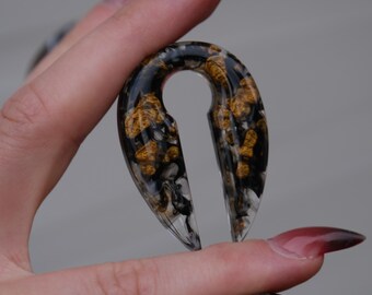 May include: Clear resin ear gauges with black and gold inclusions. The teardrop-shaped plugs have a keyhole design. The gauges are held up by a hand with long, pointed nails. The plugs are a unique accessory.