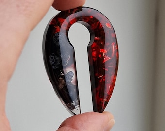 May include: A teardrop-shaped, red and black resin ear gauge with a hole in the center. The red side is filled with red glitter and the black side is filled with black glitter.