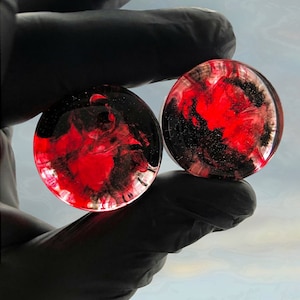 Può includere: Due tappi rotondi e trasparenti con un design a spirale rosso e nero. I tappi sono tenuti da una mano guantata, che mostra gli intricati motivi all'interno della resina. Gioielli per piercing.