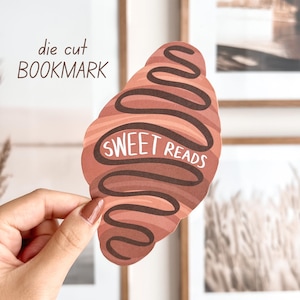 Op de afbeelding: Een gestanst boekenlegger in de vorm van een croissant, met bruine en beige tinten en de tekst "SWEET READS" in het wit. De boekenlegger wordt in de hand van een persoon gehouden.