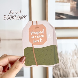 Op de afbeelding: Een gestanst bladwijzer in de vorm van een theezakje, met de tekst "steeped in a good book". De bladwijzer heeft een lichtroze achtergrond met een groen thee-achtig ontwerp aan de onderkant. De woorden "die cut BOOKMARK" staan bovenaan.