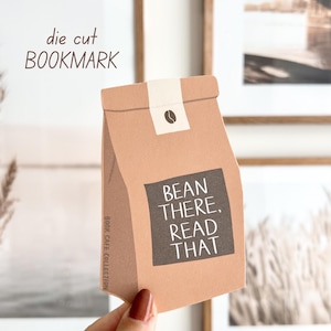 Op de afbeelding: Uitgestanste bladwijzer in de vorm van een koffiebonenzak. Het bruine papieren zakontwerp heeft de tekst "BEAN THERE, READ THAT" in een donker vierkant. De bovenkant heeft een witte band met een koffieboongrafiek.