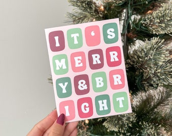 May include: A pink card with the text "It's Merry & Bright" in white letters on a green, pink, and red background. The card is being held in front of a Christmas tree.