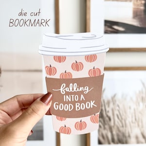 Op de afbeelding: Een uitgestoken bladwijzer in de vorm van een koffiekopje, met een patroon van oranje pompoenen op een lichtroze achtergrond. De beker heeft een bruine mouw met de tekst "falling INTO A GOOD BOOK". De bovenkant van de beker is wit.