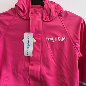 May include: A bright pink hooded raincoat featuring a front snap-button closure and a hood. The name Freya S.M. is embroidered in white on the chest. A white product tag with the text DRY KIDS is attached to the front of the jacket.