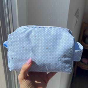May include: A light blue polka dot cosmetic bag with a zipper closure. The bag has small white polka dots and is embellished with small, colorful rhinestones. The bag has a rectangular shape with a handle on each side.