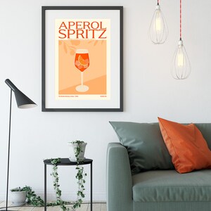 May include: A black framed poster with the text "Aperol Spritz" in red and orange letters. The poster features an illustration of a glass of Aperol Spritz with an orange slice and ice cubes.