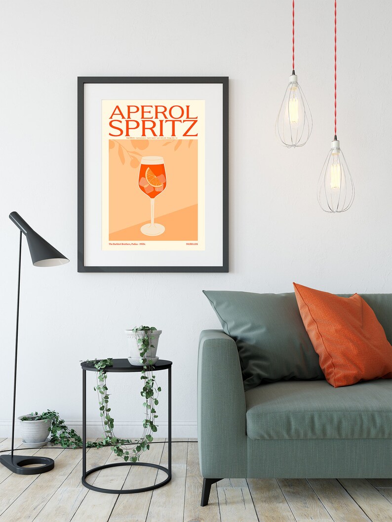 May include: A black framed poster with the text "Aperol Spritz" in red and orange letters. The poster features an illustration of a glass of Aperol Spritz with an orange slice and ice cubes.