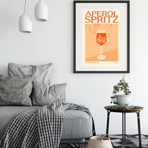 May include: A black framed poster with the text "Aperol Spritz" in red and orange letters. The poster features a glass of Aperol Spritz with an orange slice and ice cubes on a peach background.