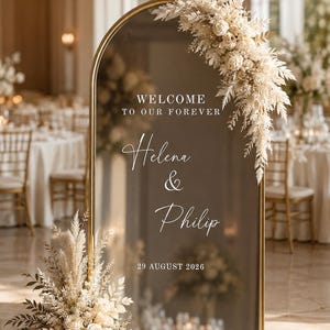 Könnte beinhalten: Ein bogenförmiges Spiegelschild mit Goldrahmen und dem weißen Text "WELCOME TO OUR FOREVER", "Helena & Philip" sowie "29 AUGUST 2026". Der Spiegel ist oben und unten mit Gestecken aus getrockneten cremefarbenen Blumen und Pampasgras verziert.
