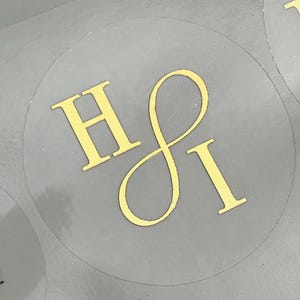 Könnte beinhalten: Klare, runde Aufkleber mit goldfarbenen Buchstaben. Die Aufkleber zeigen die Buchstaben "H" und "I" in einer stilisierten Schriftart. Die Aufkleber sind auf einer grauen Oberfläche angeordnet.