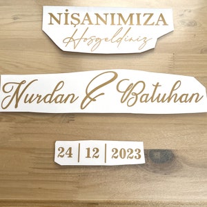 Op de afbeelding: Wit papier met gouden letters die een verloving aankondigen. De tekst luidt "Nisanımıza Hosgeldiniz" en "Nurdah & Batuhan" met de datum "24 | 12 | 2023".