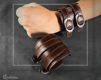 May include: Two brown leather cuffs with silver buckles. The cuffs are double layered and have a stitched design. The text "LEATHER CUFF DOUBLE STRIP" is visible at the top of the image.