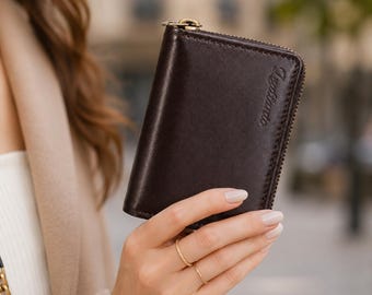 May include: A dark brown leather wallet with a smooth finish and a gold-toned metal zipper closure. The word Legtario is embossed in a script font on the front right side of the wallet. The wallet has a rectangular shape with rounded corners.