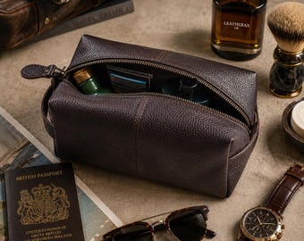 May include: A dark brown pebbled leather wash bag with a top zip fastening and a side handle. The bag is open, showing various grooming items inside. The exterior features a textured finish and visible stitching along the central panel.