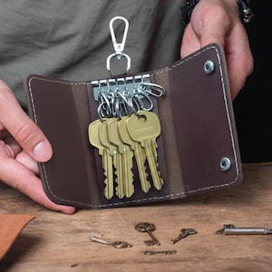 May include: A brown leather key holder with a silver clasp, holding several gold keys. The key holder is open, revealing the keys. Additional antique keys are scattered on a wooden surface.