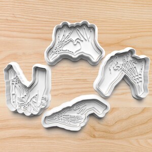 May include: Four white plastic cookie cutters shaped like hands with skeletal bones. The hands are in different poses, including a heart shape, a pinky promise, and a handshake.
