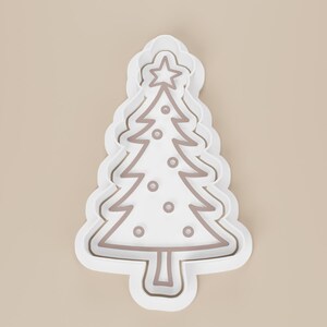 May include: A white Christmas tree cookie cutter with a brown outline and star on top. The tree has decorative circles and a scalloped edge. Perfect for holiday baking.