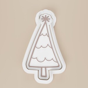 May include: A white plastic Christmas tree cookie cutter with a star on top. The tree has a wavy design and a brown outline. The cookie cutter is on a beige background.