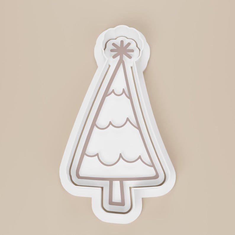 May include: A white plastic Christmas tree cookie cutter with a star on top. The tree has a wavy design and a brown outline. The cookie cutter is on a beige background.