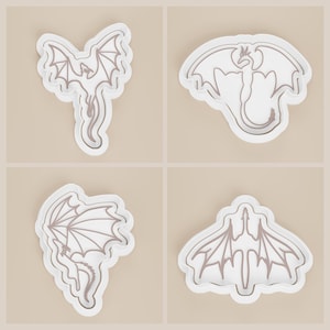 May include: Four white dragon-shaped cookie cutters with detailed wing and body designs. Each cutter has a raised outline, creating a defined shape. The cutters are displayed on a light beige background.