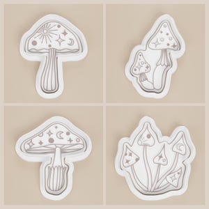 May include: Four white mushroom-shaped molds with detailed line art. Each mold features a different mushroom design, including celestial elements like stars, moons, and suns. The molds are set against a neutral background.