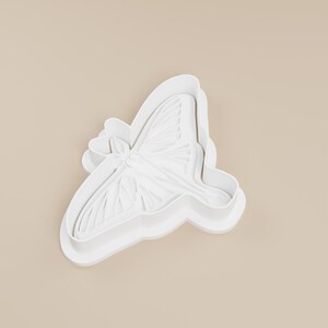 May include: A white butterfly-shaped cookie cutter. The cutter has detailed wing and body features, with a layered design. The object is made of white plastic and is set against a neutral background.