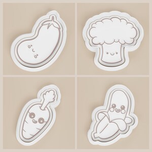 May include: Four white vegetable-shaped cookie cutters with smiling faces. The set includes an eggplant, broccoli, carrot, and banana. Each cutter has a light brown outline and is set against a beige background.