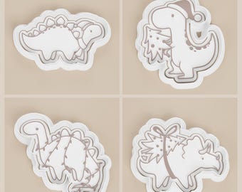 Pode incluir: Quatro cortadores de biscoitos brancos em forma de dinossauro. Um é um Estegossauro com um laço, outro um T-Rex usando um gorro de Papai Noel e segurando uma árvore de Natal. O terceiro tem uma corda de luzes e o quarto uma árvore de Natal com um laço.