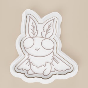 May include: A white cookie cutter in the shape of a cartoon moth with large eyes, feathery antennae, and a fluffy body. The moth is outlined in a light brown color, and the cutter has a raised edge.