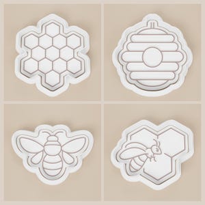 May include: Four white and gray cookie cutters with a bee and honeycomb theme. The set includes a honeycomb, a beehive, a bee, and a bee on a honeycomb. These cutters are ideal for making themed cookies or other baked goods.
