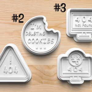 May include: Four white plastic cookie cutters shaped like computer screens and a cookie. The cookie cutter with the cookie has the text "I'M DELETING COOKIES". The other cookie cutters have the text "404" and "Not Found".