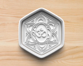 May include: White plastic cookie cutter shaped like a hexagon with an intricate mandala design featuring the Om symbol in the center.