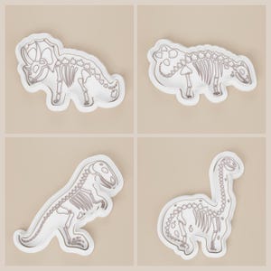 May include: Four white dinosaur-shaped cookie cutters with gray skeletal designs. The set includes a Triceratops, Ankylosaurus, Tyrannosaurus Rex, and Brachiosaurus. Each cutter has a white border and is set against a beige background.
