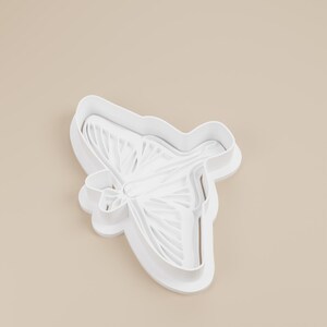May include: A white plastic cookie cutter in the shape of a dragonfly. The cutter has detailed wing and body features, with a smooth, clean finish. The dragonfly design is suitable for baking and crafting.