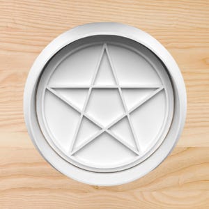 May include: A white, circular cookie cutter featuring a pentagram design. The cutter has a raised edge and a detailed star shape within a circle. The object is made of white plastic and sits on a light wood surface.