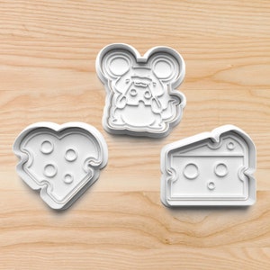 May include: Three white plastic cookie cutters shaped like a mouse holding a piece of cheese, a heart-shaped piece of cheese, and a wedge of cheese.