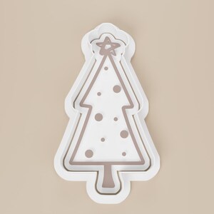 May include: A white Christmas tree-shaped cookie cutter with a star on top and decorative dots. The cutter has a layered design with a light brown interior and a white exterior.