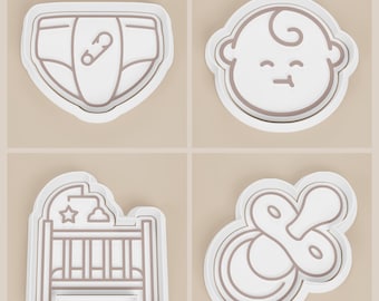 May include: Four white and brown baby-themed cookie cutters. The shapes include a diaper with a safety pin, a baby's face, a crib with a mobile, and a pacifier. These cutters are ideal for creating baby shower treats.