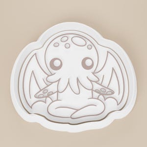 May include: A white cookie cutter in the shape of a cartoon Cthulhu. The design features a stylized creature with large eyes, tentacles, and wings. The cutter has a rounded, cloud-like border.