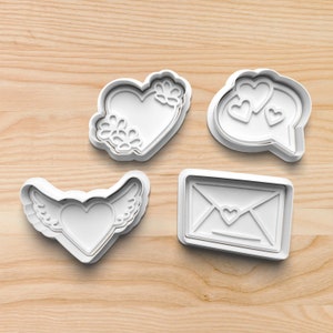 May include: Four white plastic cookie cutters shaped like a heart with wings, a heart with flowers, a speech bubble with hearts, and an envelope with a heart.