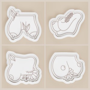 May include: Four white cookie cutters with a light brown outline. The cutters are in the shape of stylized body parts, including breasts and buttocks, with floral and leaf accents. The background is a solid light beige.