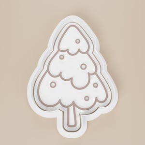 May include: A white plastic Christmas tree-shaped cookie cutter with a light brown outline. The tree has three tiers with rounded edges and small circular details. The cutter is designed for baking and crafting.
