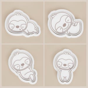 May include: Four white sloth-shaped cookie cutters with gray outlines. Each cutter depicts a different pose of a cartoon sloth. The sloths have closed eyes and are smiling. The cutters are on a light beige background.